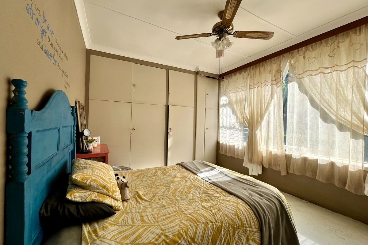 A bedroom featuring built-in cupboards, a ceiling fan, and large windows with security bars, offering a functional space with natural light.