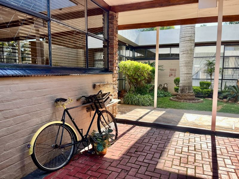 A covered patio area featuring brick paving, exposed brick walls, and large steel-framed windows overlooking a landscaped courtyard garden.