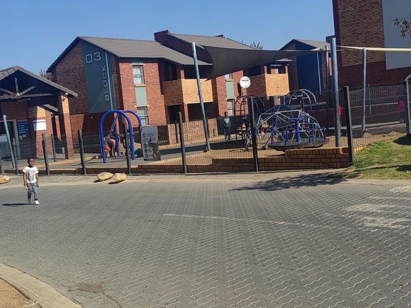 Exterior view of an apartment complex featuring brick architecture, a paved driveway, and communal playground facilities with shade structures.