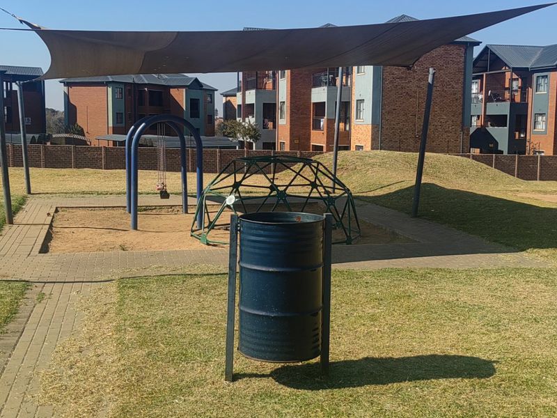Communal garden area featuring a playground with shade sail, paved pathways, and perimeter brick walling within the apartment complex.