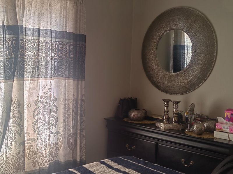 A bedroom interior featuring a window with patterned curtains and a wall-mounted decorative mirror.