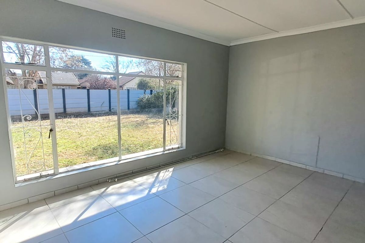 A bright, empty living room featuring large steel-framed windows with security bars, neutral tiled flooring, and coved ceilings, overlooking the rear garden.