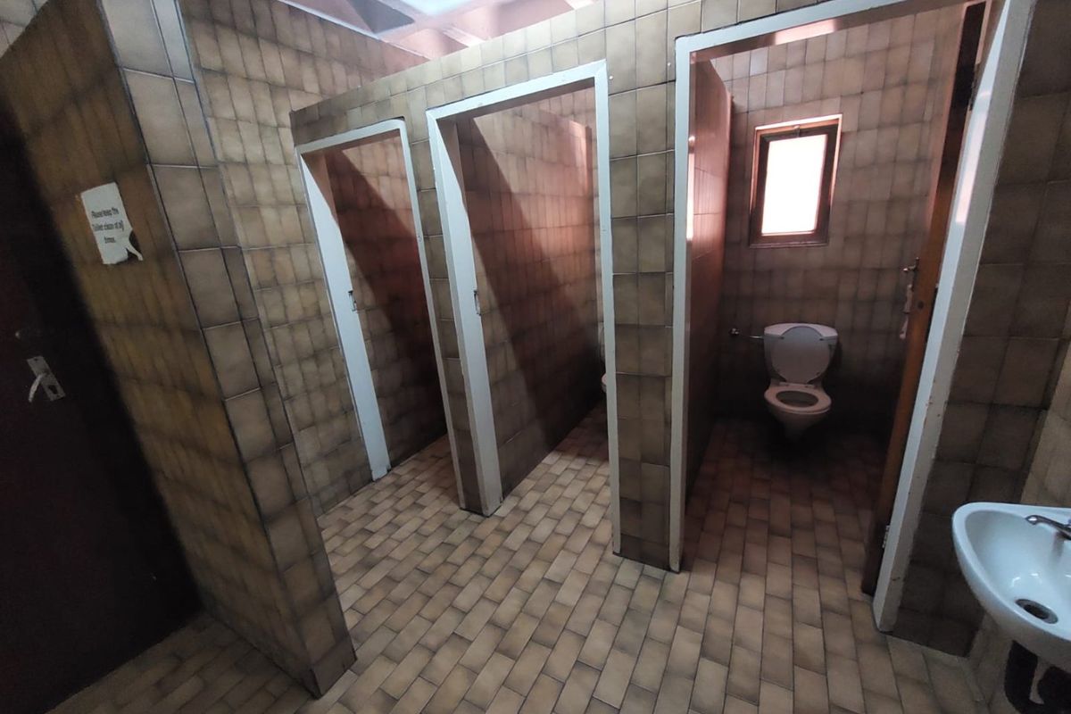 A communal office restroom featuring multiple toilet cubicles with dated ceramic wall and floor tiling and a wall-mounted basin.