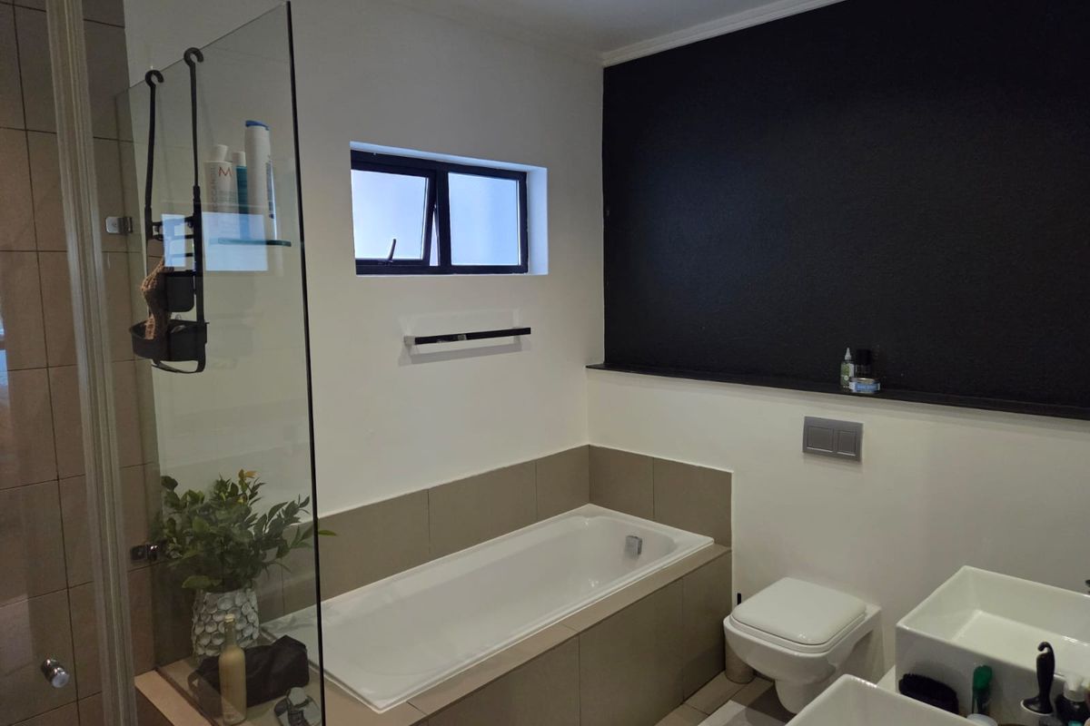 Modern bathroom featuring a built-in bathtub, wall-mounted toilet, dual vanity basins, and a glass shower screen with neutral tiling and a feature wall.