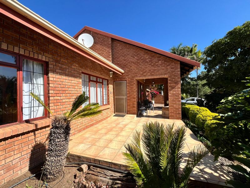 A well-maintained brick-faced patio area featuring tiled flooring, aluminum window frames, and a secure entrance, set within a landscaped garden environment.