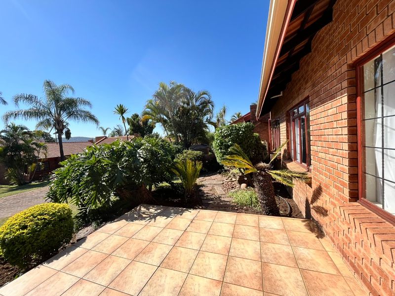 A paved outdoor patio area adjacent to the brick exterior of the house, featuring established garden landscaping and clear views of the surrounding property.