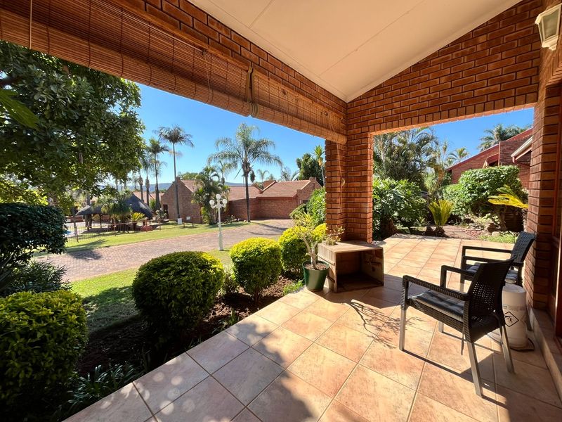 A covered patio featuring exposed brick pillars and tiled flooring, overlooking a landscaped garden and paved driveway area.