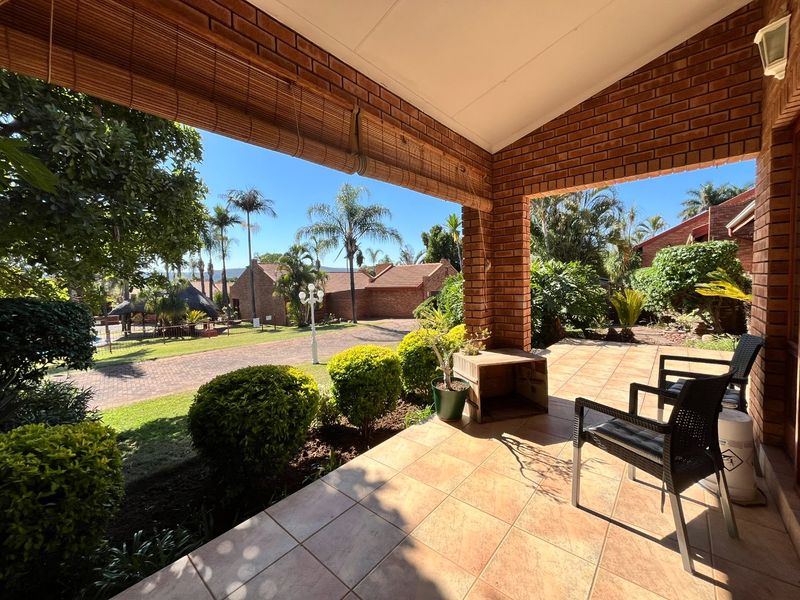A covered patio featuring exposed brick pillars and tiled flooring, overlooking a landscaped garden and paved driveway area.