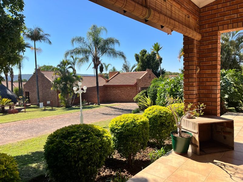 A covered patio area with tiled flooring and brick pillars, overlooking a well-maintained garden and paved driveway in a residential estate.