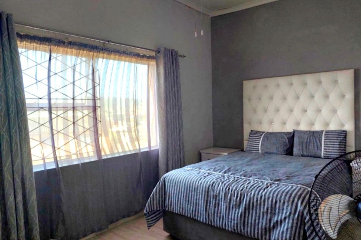 A bedroom featuring laminate flooring, a ceiling fan, and a large window with curtain rails, providing a functional and comfortable sleeping space.