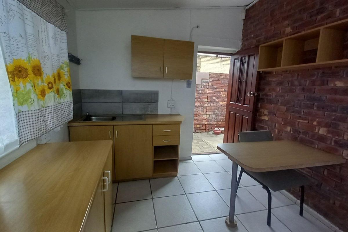 A compact kitchen area featuring light wood cabinetry, a stainless steel sink, tiled flooring, and an exposed brick feature wall with direct outdoor access.