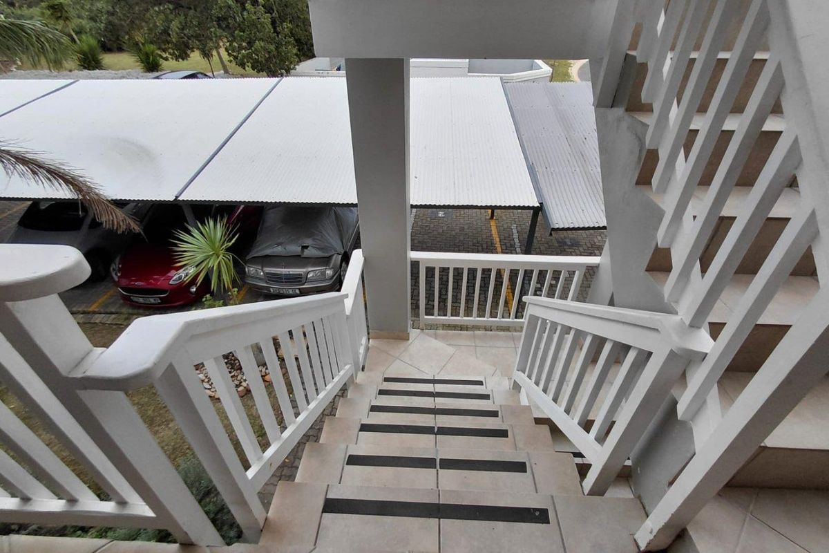An exterior view looking down a tiled staircase with white railings, overlooking a paved parking area with covered carports.