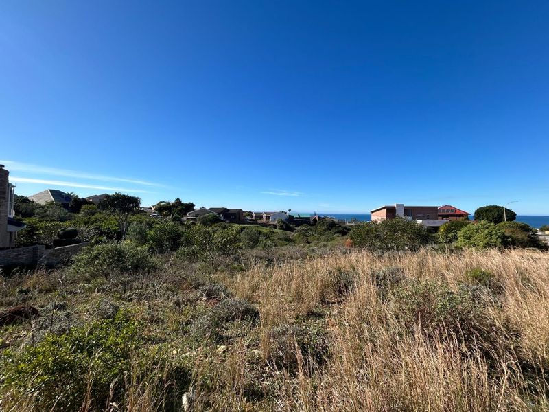 A wide-angle view of a vacant residential plot featuring natural coastal vegetation with distant ocean views and surrounding neighborhood homes.
