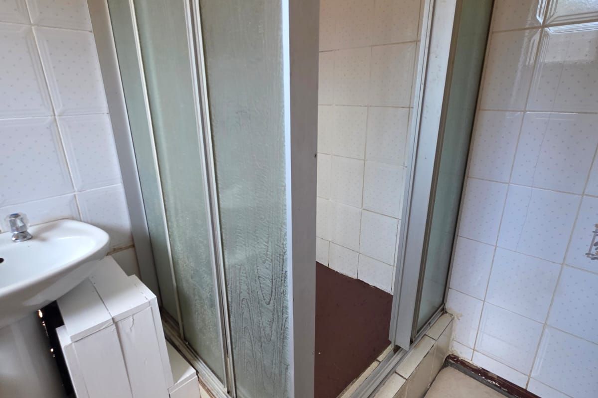 A dated bathroom featuring a pedestal sink and a shower enclosure with tiled walls and flooring, requiring modernization and cosmetic updates.