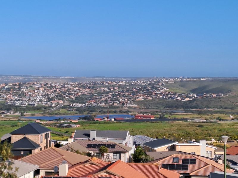 An elevated panoramic view overlooking a residential neighborhood, green landscape, and distant coastal terrain under a clear blue sky.