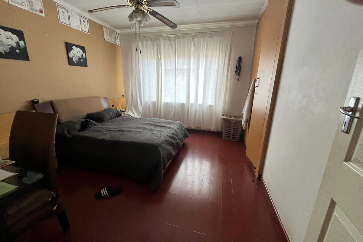 A bedroom featuring a ceiling fan, built-in wooden wardrobe, and large window with curtains, finished with painted wooden flooring.