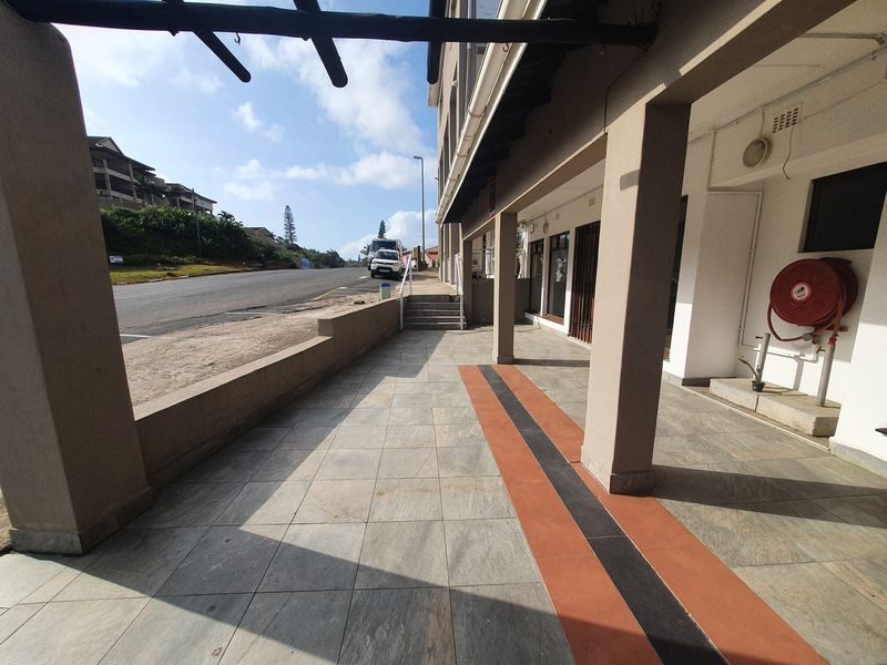 A covered outdoor walkway or patio area featuring tiled flooring, concrete support pillars, and a fire hose reel, adjacent to a street-facing commercial facade.
