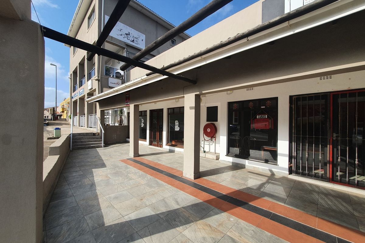 A commercial exterior walkway featuring tiled flooring, a pergola structure, and secure glass storefronts with metal security gates.