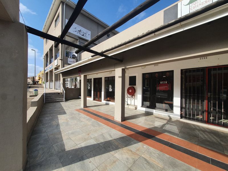A commercial exterior walkway featuring tiled flooring, a pergola structure, and secure glass storefronts with metal security gates.