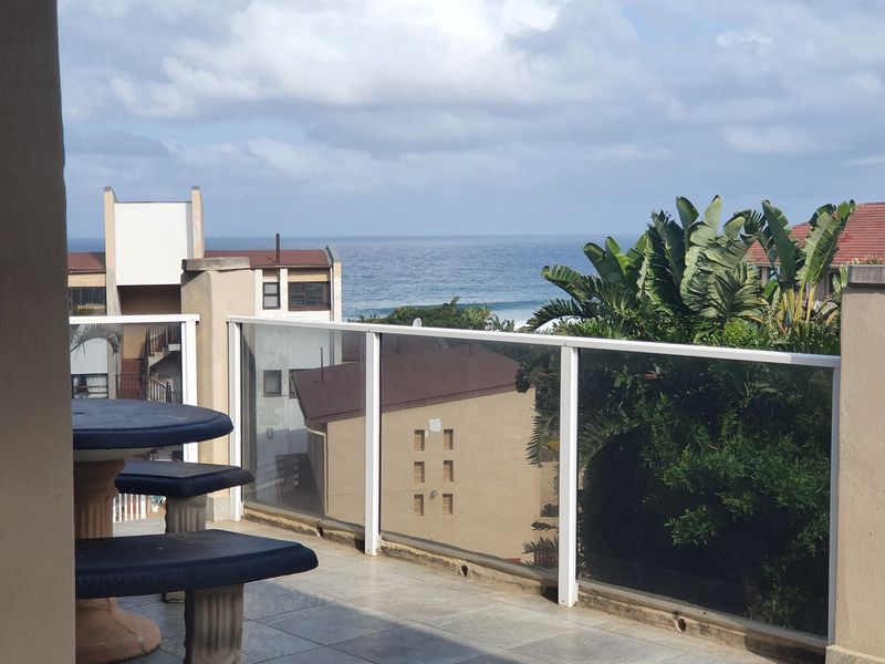 A balcony featuring a glass balustrade and tiled flooring, offering an elevated view of the surrounding buildings and the ocean.