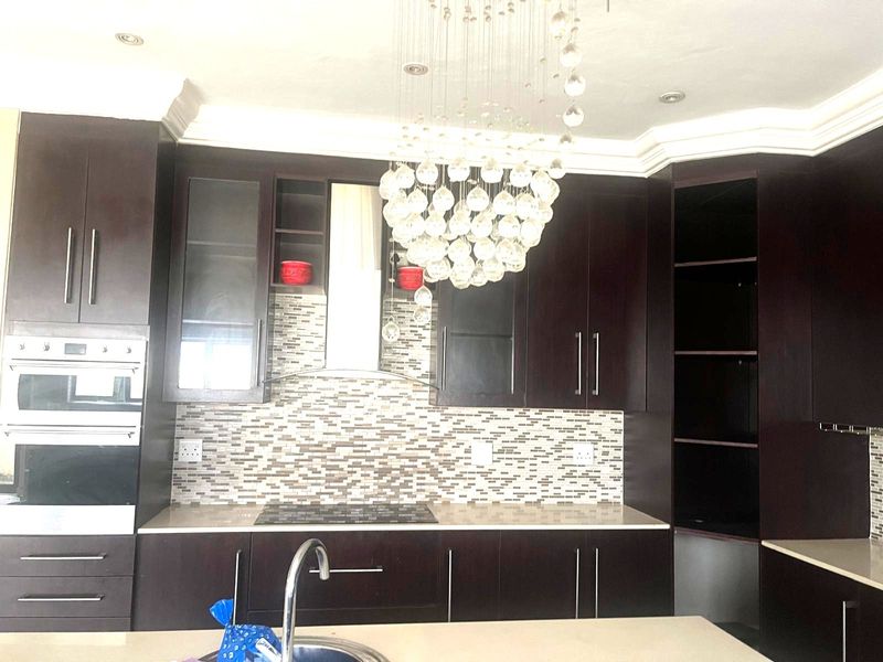 Modern kitchen featuring dark cabinetry, stone countertops, a mosaic tile backsplash, built-in oven, and a prominent decorative chandelier.