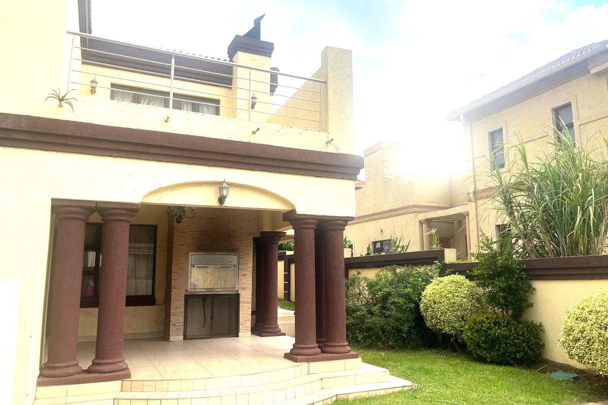 Rear exterior view featuring a covered patio with a built-in braai, tiled flooring, decorative columns, and an upper-level balcony with stainless steel railings.