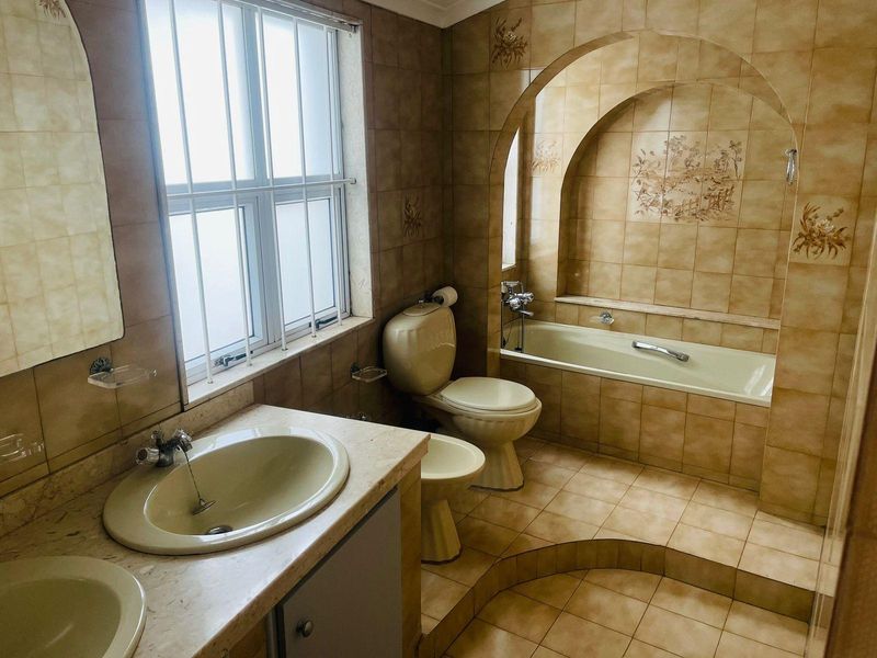 A spacious bathroom featuring a built-in bathtub with an arched alcove, dual vanity basins, a toilet, and a bidet, finished with dated ceramic tiling.