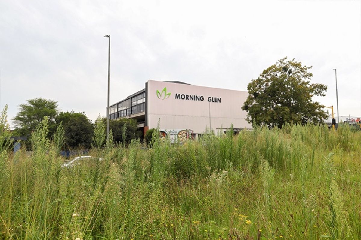 A view of a vacant residential plot with overgrown grass, featuring a commercial building in the background.