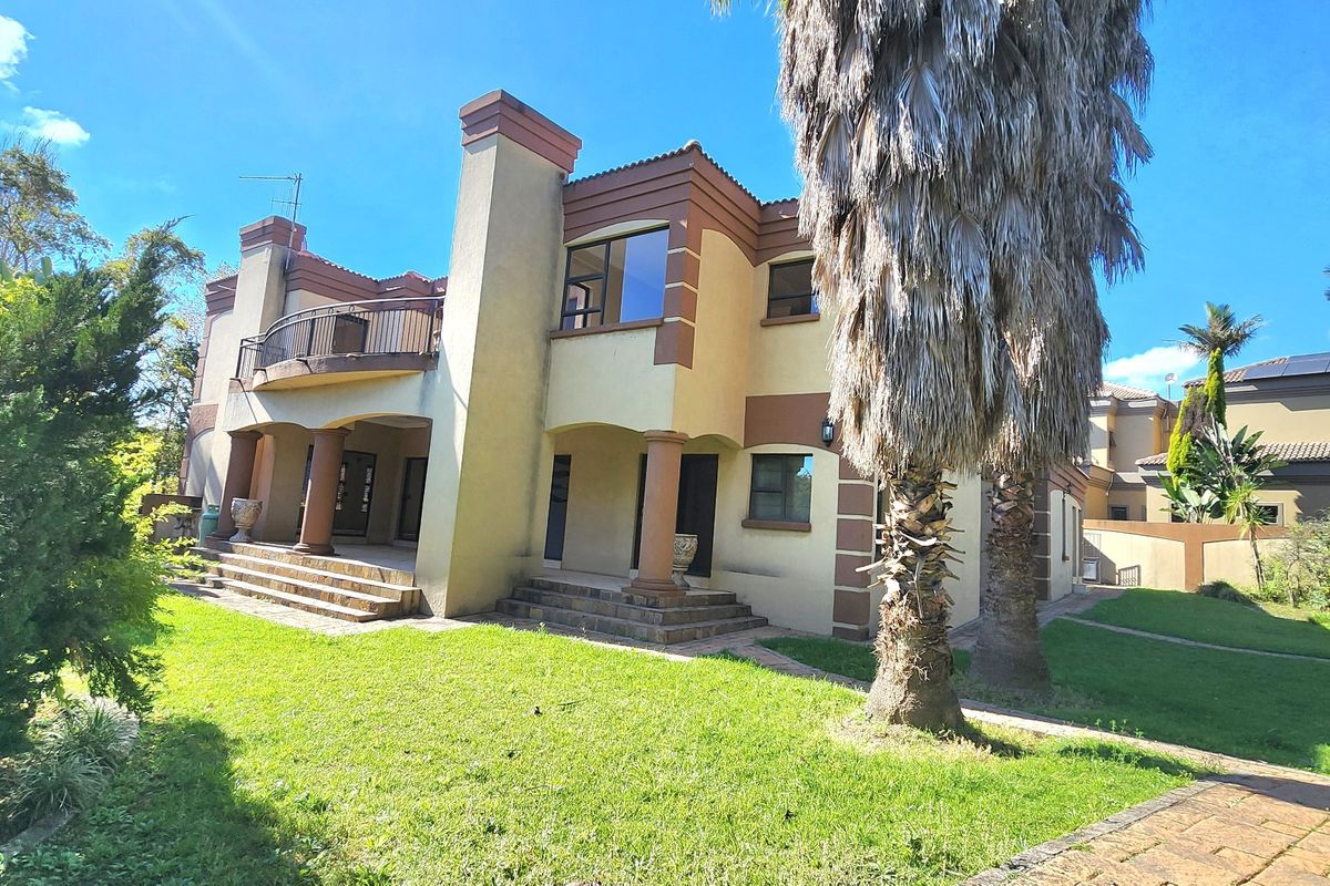 Rear view of a multi-story house featuring a balcony, stone columns, a chimney, and a spacious garden with paved patio areas.