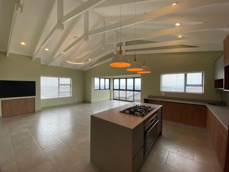 Spacious open-plan living and kitchen area featuring a vaulted ceiling with exposed rafters, a central island with built-in appliances, and large windows with views.