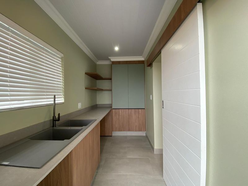 A modern, well-maintained scullery featuring double sinks, sleek wood-finish cabinetry, floating shelves, and a sliding door, providing efficient additional prep and wash-up space.