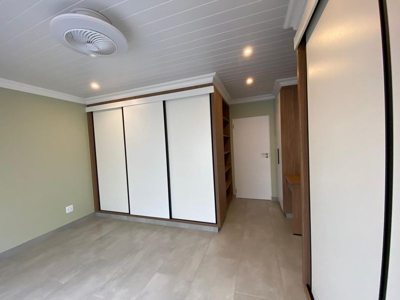A modern, empty bedroom featuring built-in sliding door cupboards, tiled flooring, a ceiling fan, and recessed lighting with a clean, neutral finish.