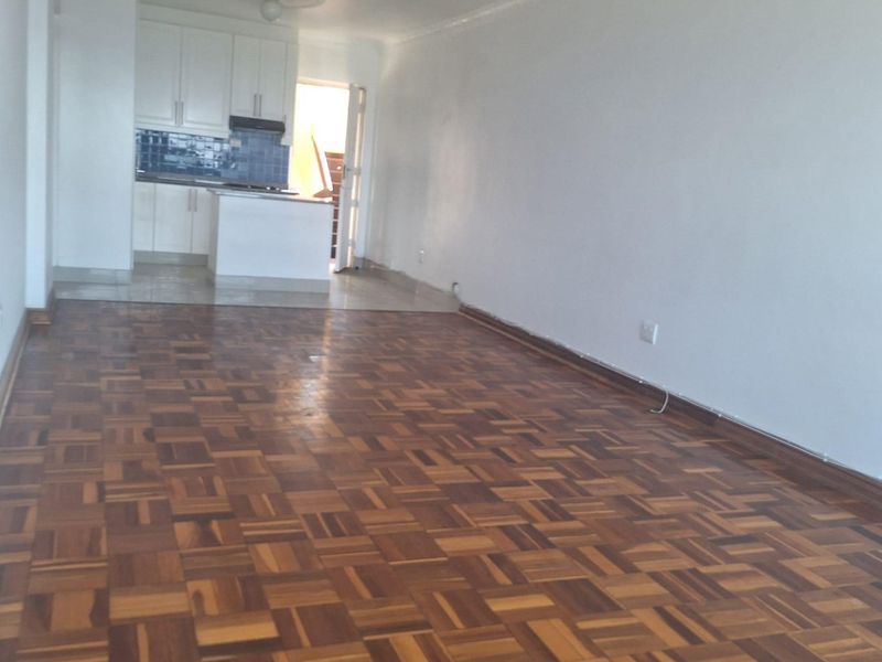An open-plan living and kitchen area featuring classic parquet flooring, white cabinetry, and a tiled splashback in a vacant apartment setting.