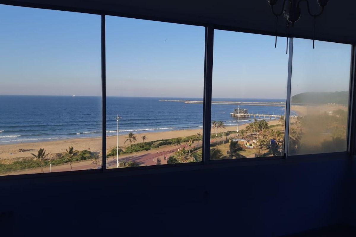 A clear, elevated ocean and beach view captured through large, fixed-pane windows in an apartment.