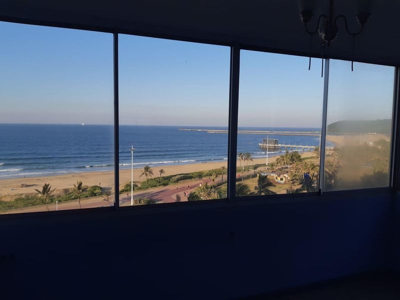 A clear, elevated ocean and beach view captured through large, fixed-pane windows in an apartment.