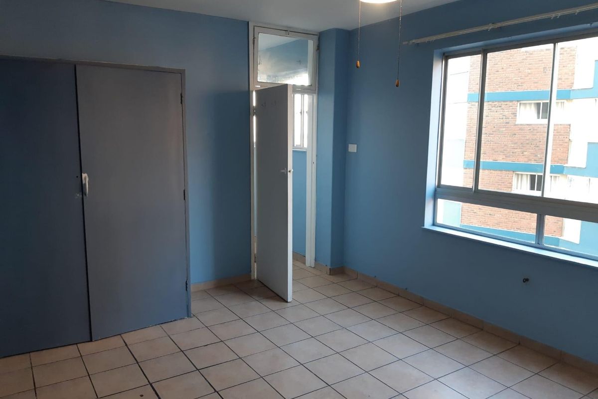 An empty bedroom featuring blue walls, tiled flooring, a built-in cupboard, and a large window providing natural light.