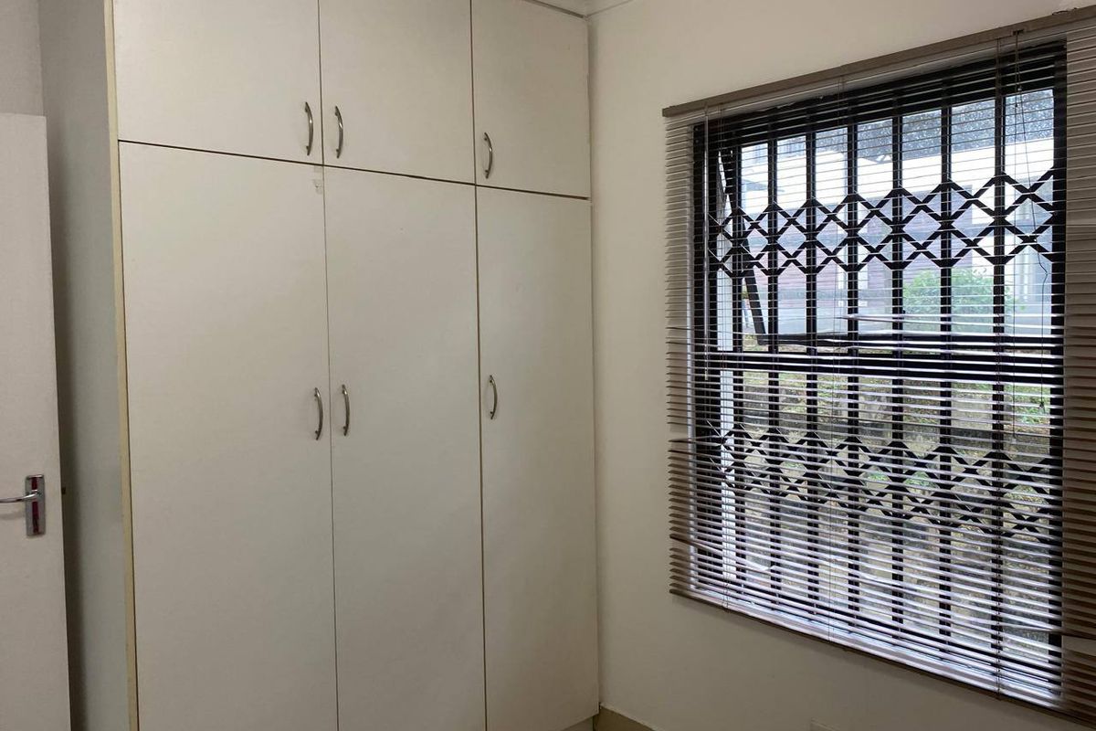A clean, empty bedroom featuring white built-in cupboards, tiled flooring, and a window fitted with a security gate and venetian blinds.