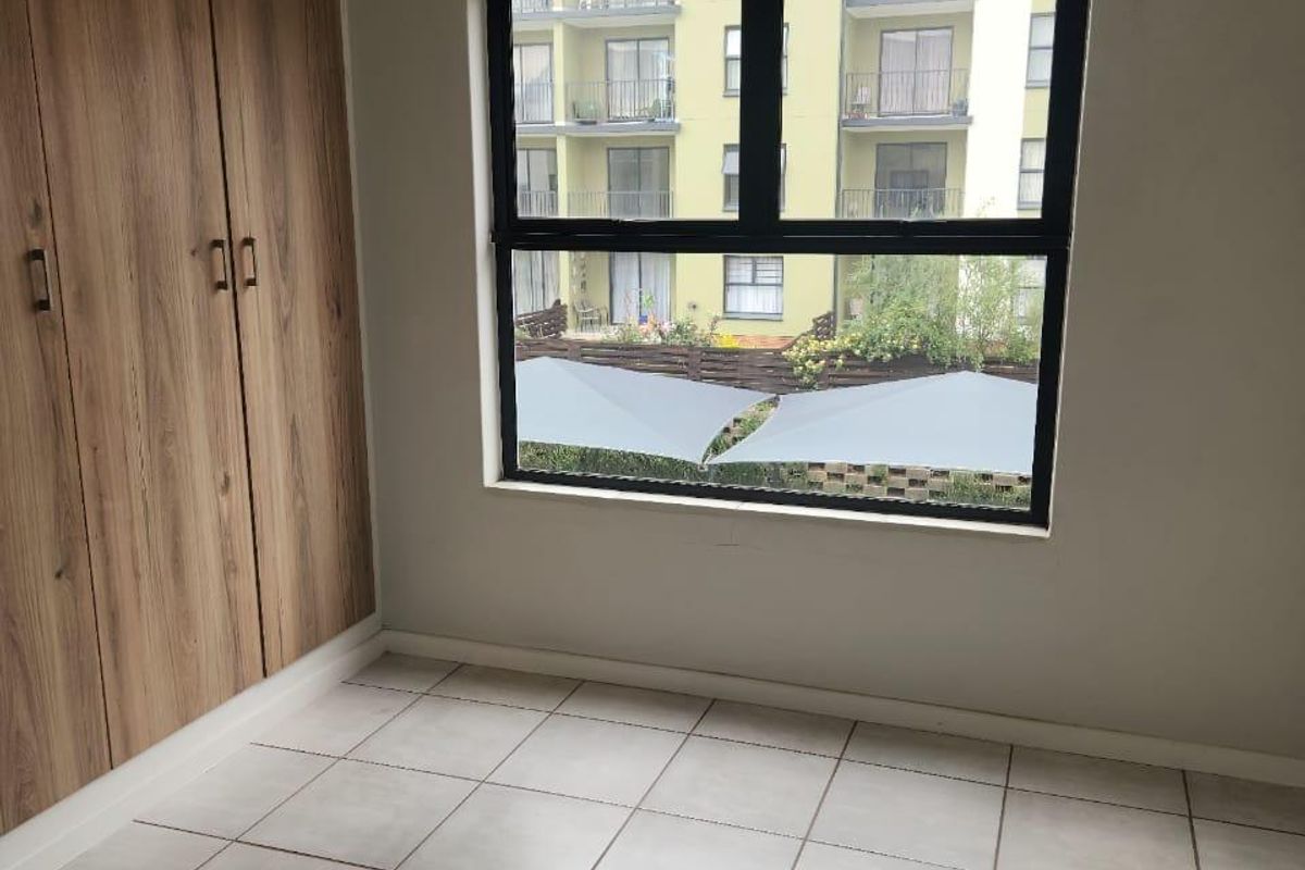 A bright, empty bedroom featuring neutral tiled flooring and modern wood-finish built-in cupboards with a view of the surrounding apartment complex.