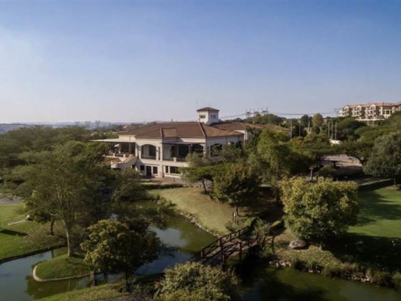 Aerial view of a residential property featuring landscaped grounds, a water feature, and a pedestrian bridge leading to the main building.