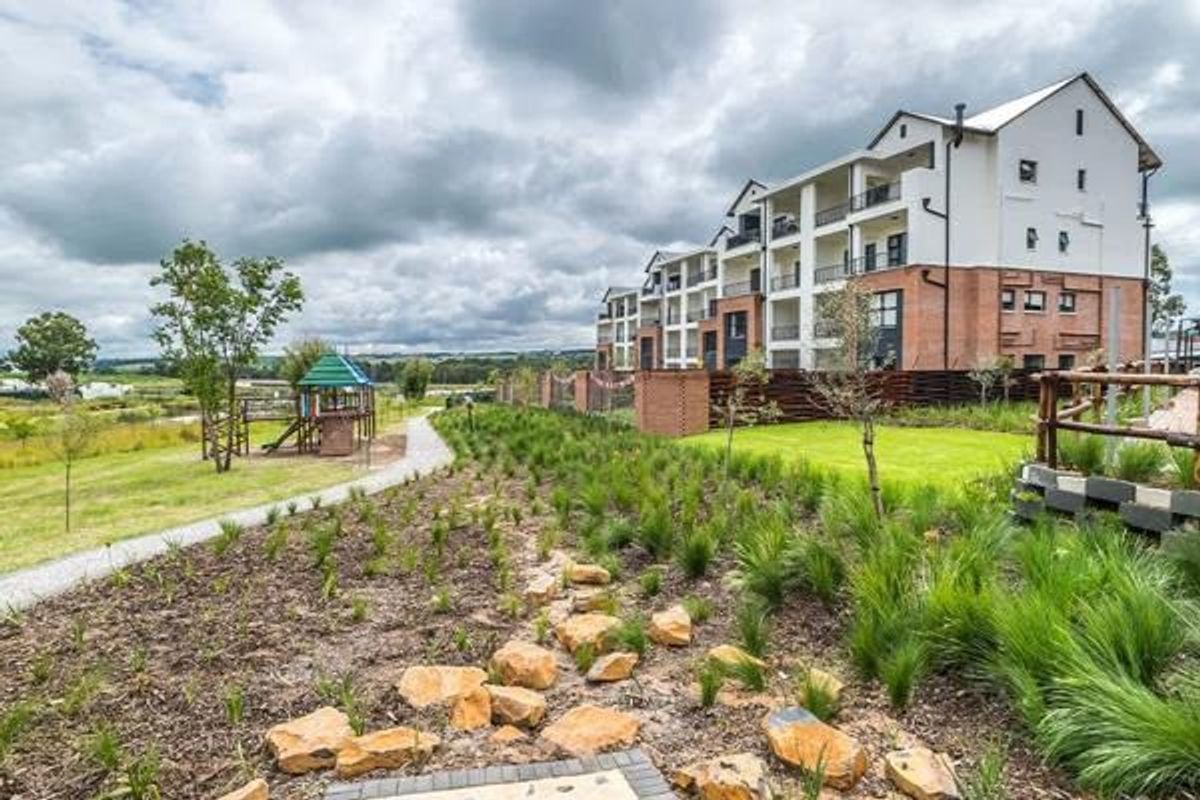 A view of the apartment complex exterior featuring landscaped gardens, a paved walkway, and a communal playground area.