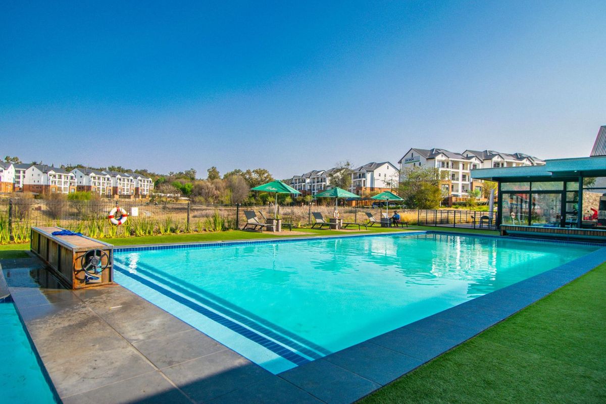 A well-maintained outdoor swimming pool area featuring modern tiling, artificial turf surrounds, and adjacent clubhouse facilities within a residential apartment complex.