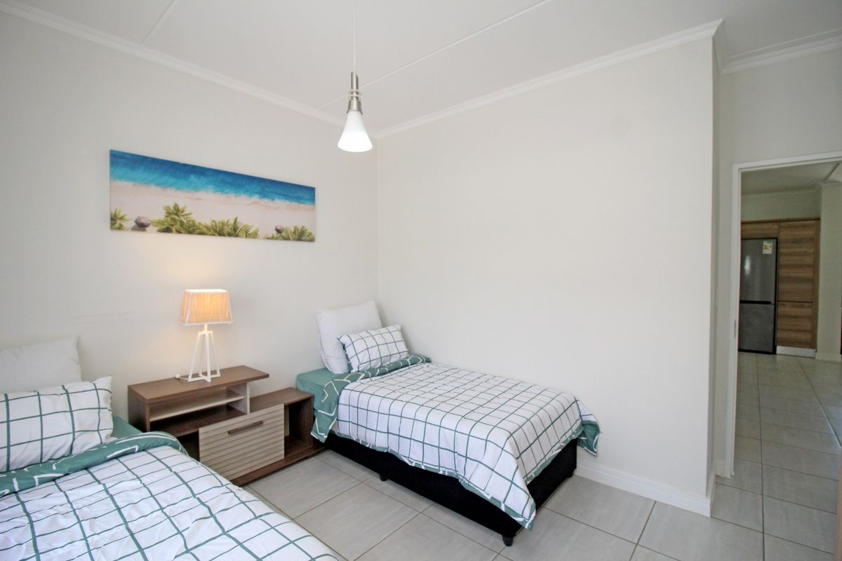 A clean, neutrally decorated bedroom featuring tiled flooring, coved ceilings, and a pendant light fixture, with an adjacent view into the kitchen area.
