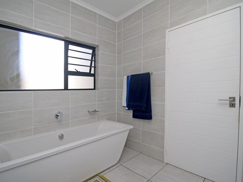 A modern bathroom featuring neutral grey wall and floor tiles, a clean white freestanding bathtub, and a frosted glass window for privacy.