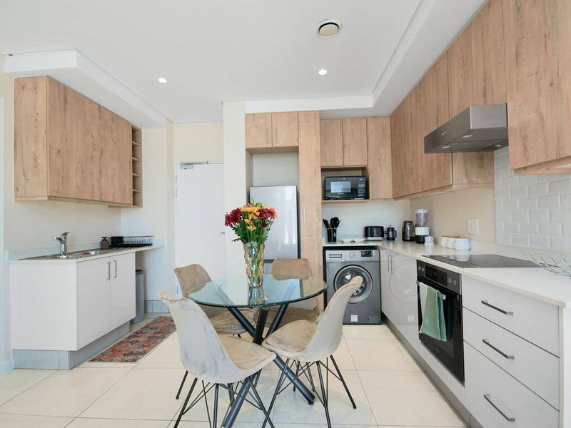 Modern open-plan kitchen featuring light wood-tone cabinetry, stone countertops, subway tile backsplash, and integrated appliances including a washing machine and oven.
