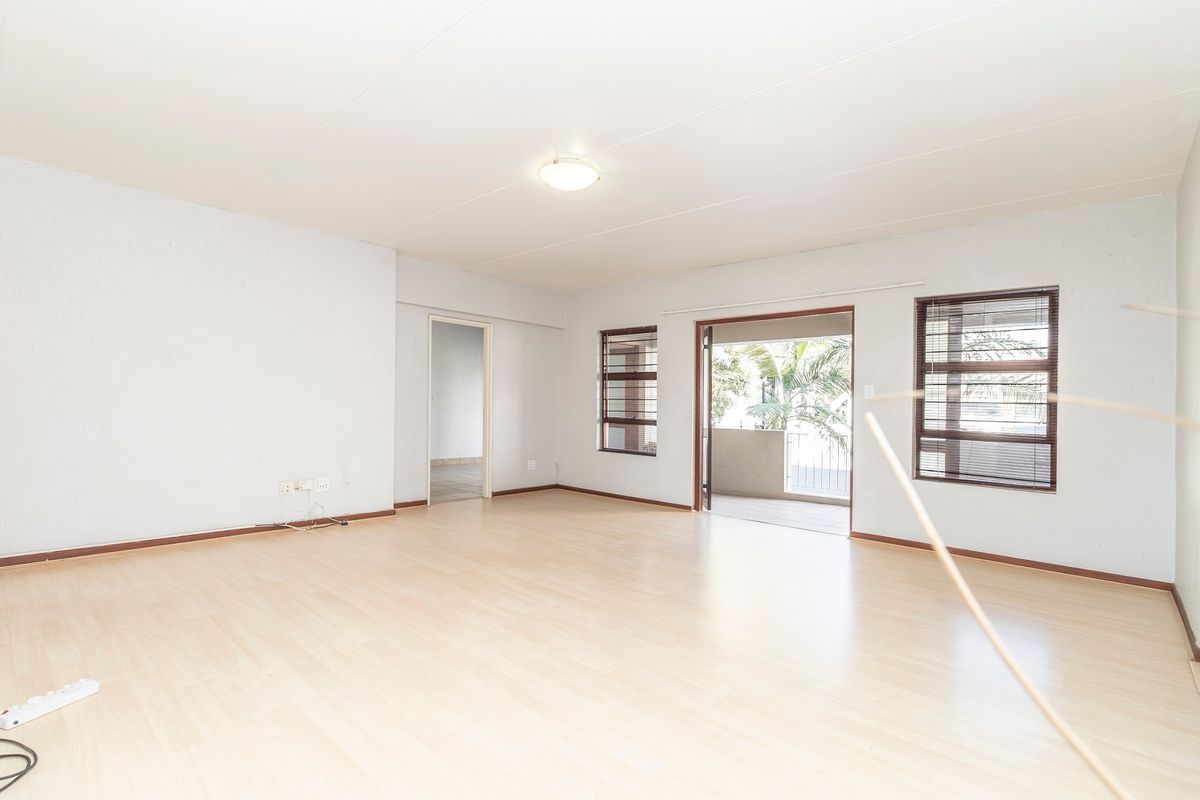 A bright, empty living room featuring light-toned laminate flooring, large windows with venetian blinds, and a sliding door leading to an outdoor balcony.