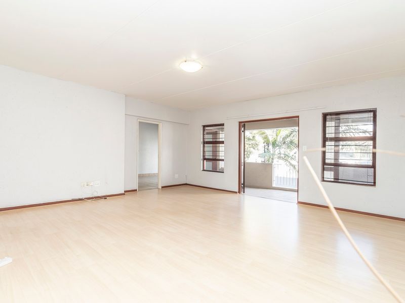 A bright, empty living room featuring light-toned laminate flooring, large windows with venetian blinds, and a sliding door leading to an outdoor balcony.