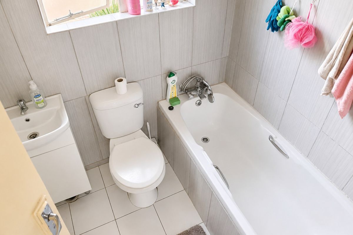 A compact bathroom featuring a built-in bathtub, toilet, and vanity basin with neutral tiled finishes and natural light from a casement window.