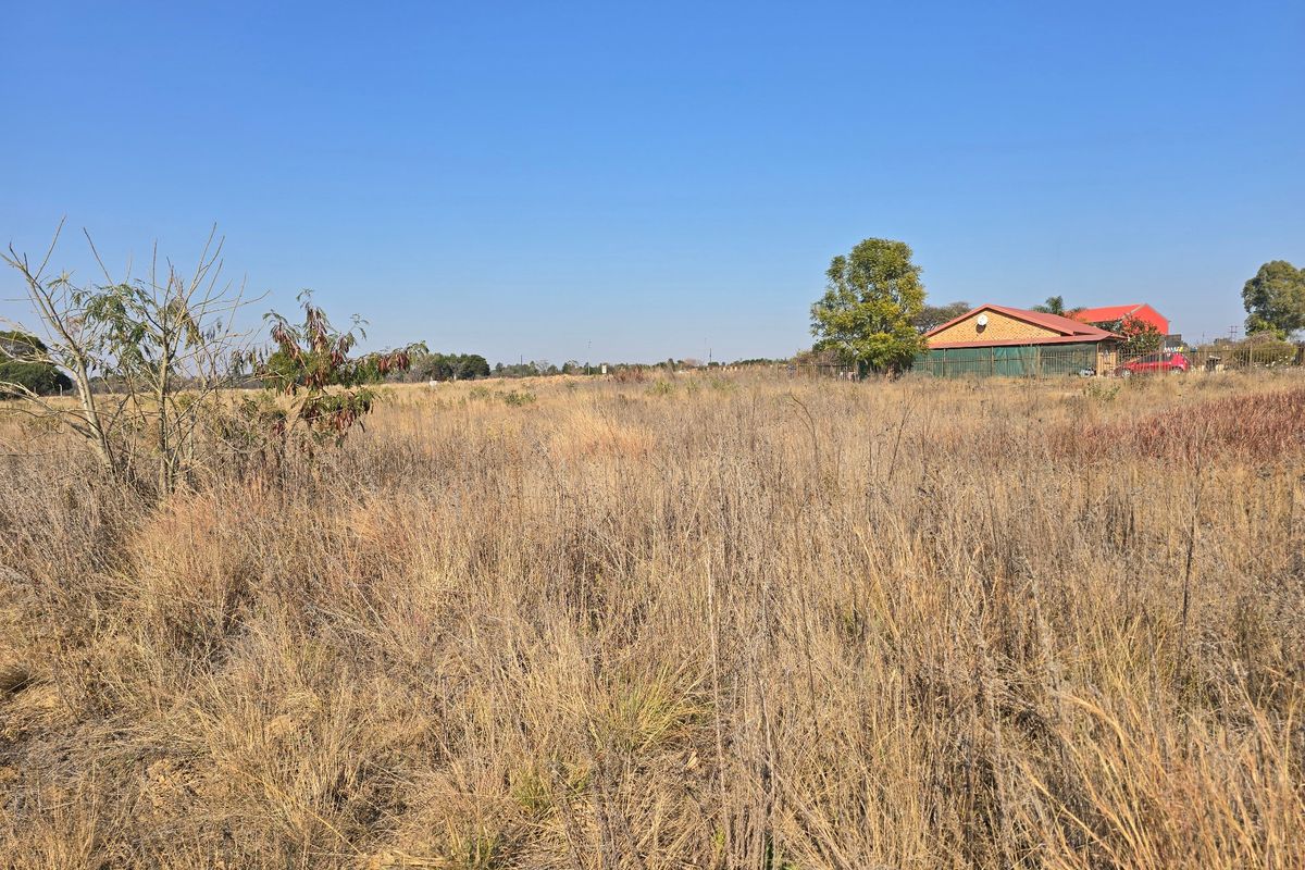 A wide-angle view of a vacant residential plot featuring natural dry grass and vegetation, with neighboring structures visible in the distance.