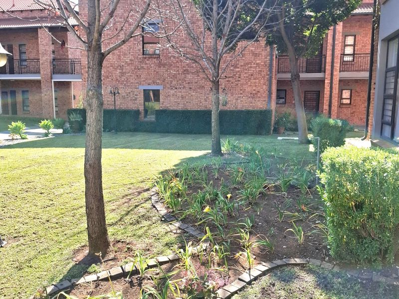 A view of the communal garden area featuring landscaped beds with brick edging, mature trees, and the brick facade of the apartment complex.