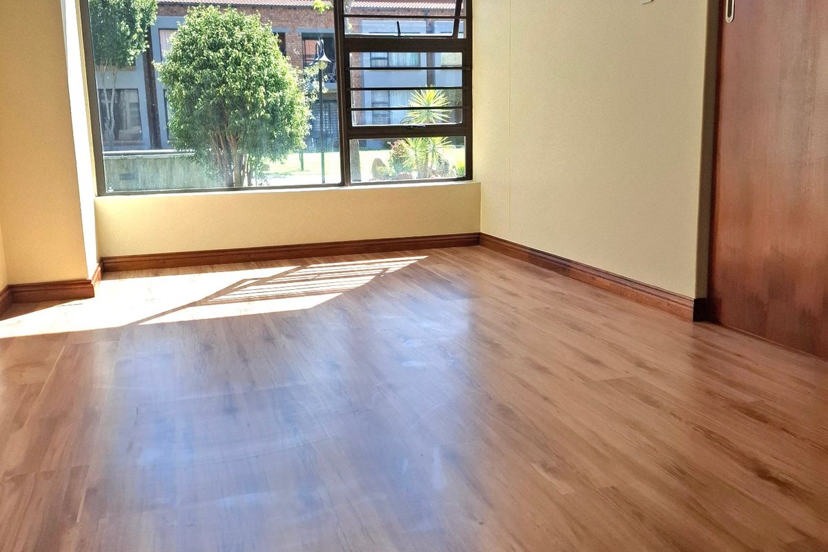 A bright and empty bedroom featuring laminate flooring, a built-in cupboard, and a large window providing natural light and a view of the complex.